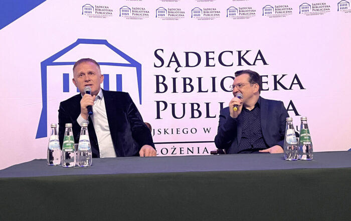 Dwóch mężczyzn siedzi za długim stołem nakrytym ciemnozielonym materiałem. Każdy z mężczyzn trzyma w ręce mikrofon blisko ust. Mężczyzna po prawej stronie patrzy na mężczyznę siedzącego obok. Na stole znajduje się pięć szklanych butelek z wodą. W tle ścianka reklamowa Sądeckiej Biblioteki Publicznej.