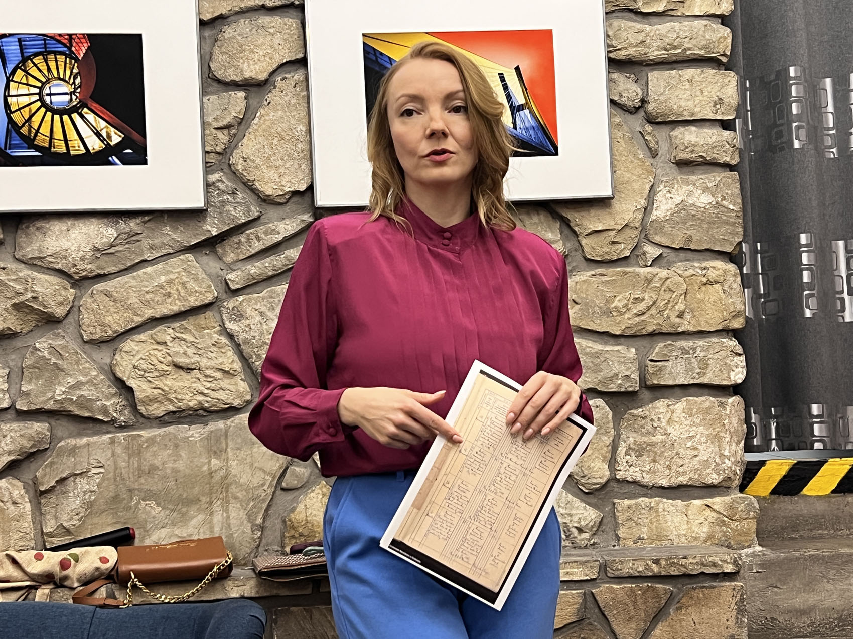 Warsztaty_Aneta Godynia (1) W pomieszczeniu o kamiennej ścianie stoi młoda kobieta ubrana w bordową bluzkę i niebieskie spodnie. W rękach trzyma kartkę z wydrukowaną tabelą. W tle na ścianie wiszą dwie kolorowe fotografie.