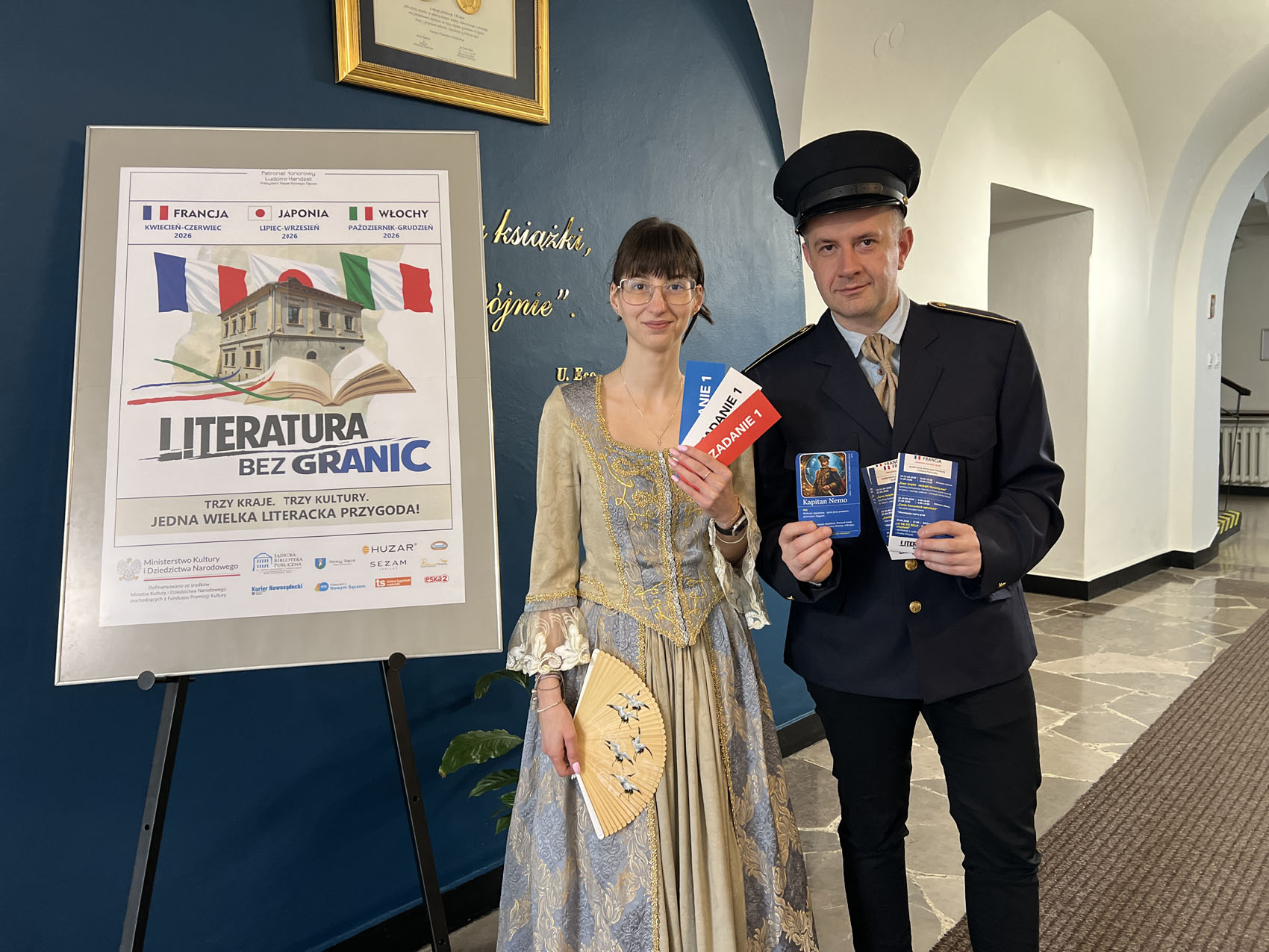 W długim korytarzu na tle niebiesko-białej ściany stoi młoda kobieta i mężczyzna. Uśmiechają się. Kobieta ubrana jest w długą, elegancką suknię. Mężczyzna ubrany jest w ciemne spodnie, granatową marynarkę i ciemną czapkę. Obydwoje trzymają w rękach ulotki, karteczki, karty gry. Po lewej stronie na czarnej sztaludze stoi obraz z grafiką.