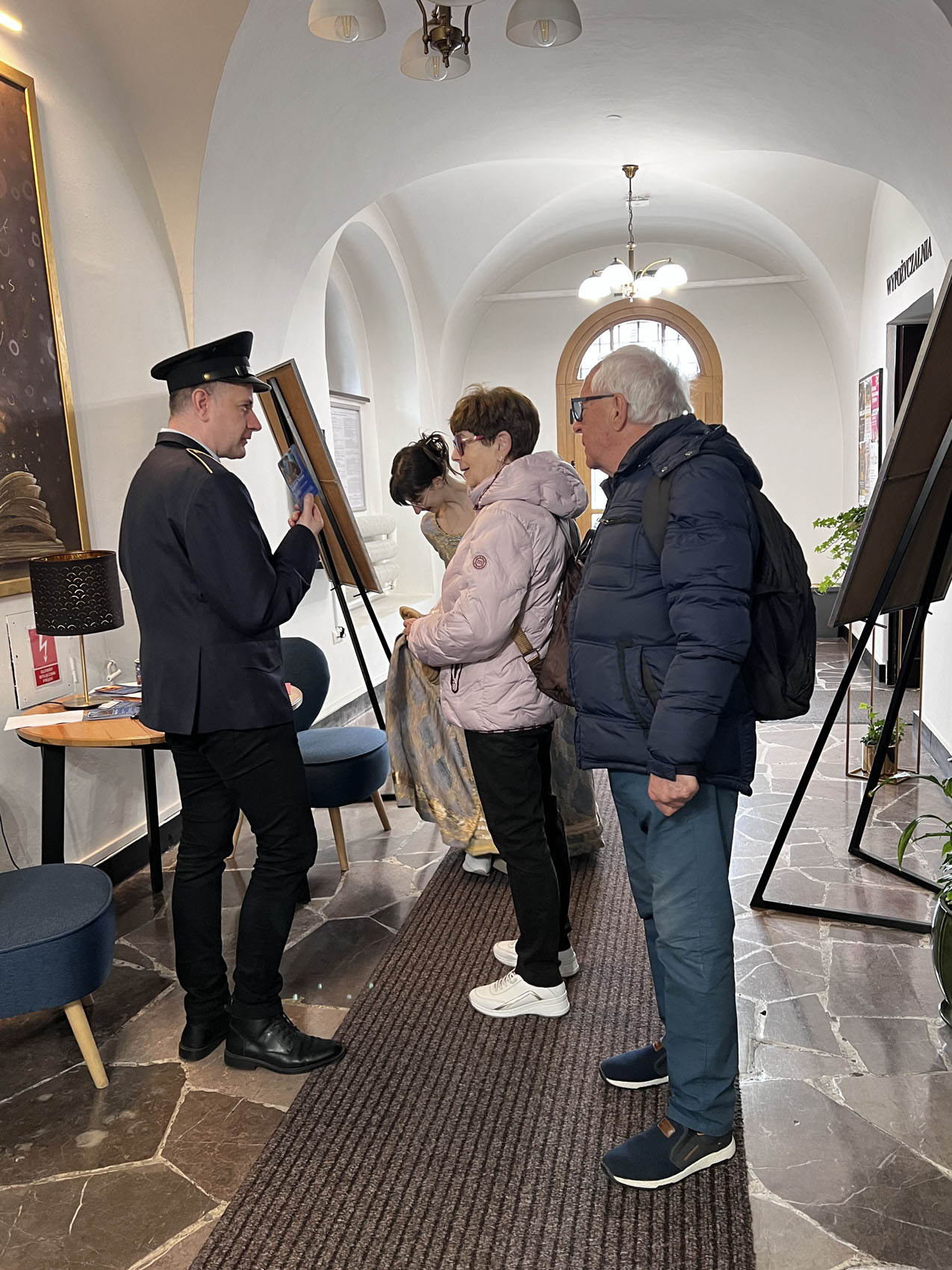 Żywe ksiażki_6 W długim korytarzu stoją cztery osoby: młody mężczyzna, starszy mężczyzna, młoda dziewczyna i kobieta. Kobieta i starszy mężczyzna stoją przed młodym mężczyzną ubranym w ciemny garnitur i czapkę. Mężczyzna trzyma w ręce karteczkę, pokazuje osobom.