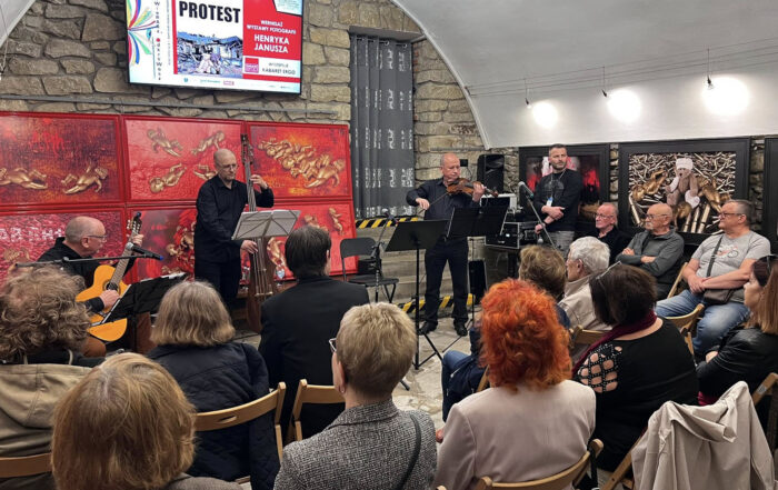 W pomieszczeniu o kamiennych ścianach i kamiennej podłodze siedzi na krzesłach grupa ludzi, pokazanych od tyłu. Przed nimi występuje zespół - trzech mężczyzn. Mężczyzna po lewej stronie gra na gitarze, mężczyzna w środku gra na kontrabasie a mężczyzna po prawej gra na skrzypcach. Na ścianach wiszą kolorowe obrazy.