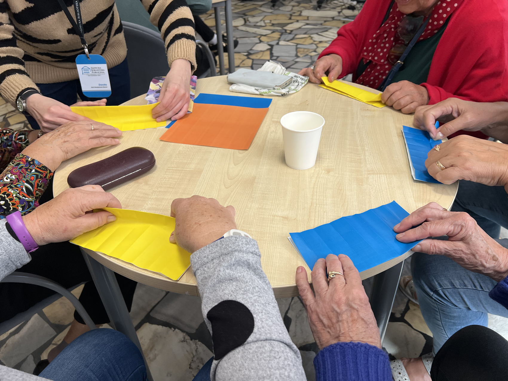 Warsztaty japońskie z seniorami (5) Grupa seniorów składa kolorowe kartki papieru w origami przy stoliku przy pomocy pracownika biblioteka
